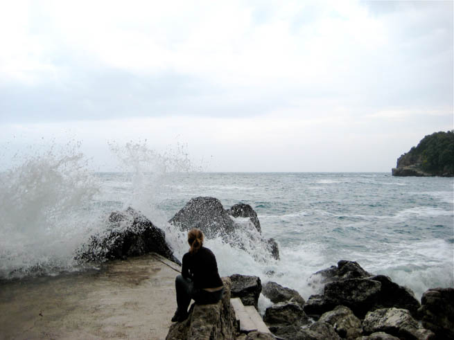 Woman watches waves crash on rocky shore in Iceland.