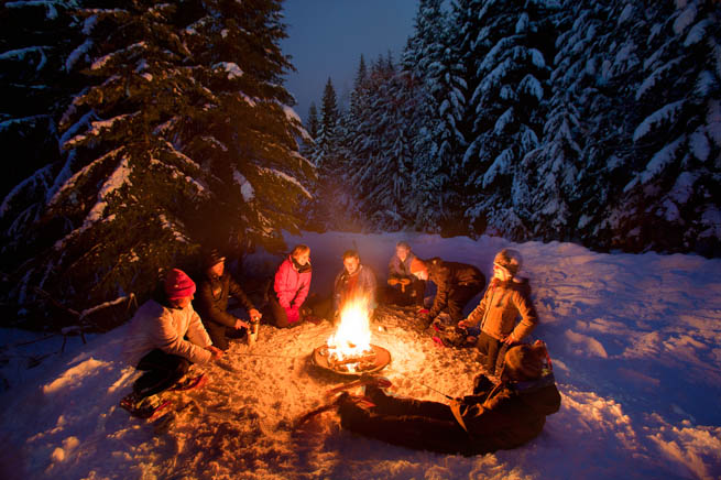 A group of people gather around a fire pit during a snowy evening in Vermont.